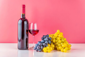 Fototapeta premium A bottle of red wine stands next to a glass filled with wine, accompanied by clusters of yellow and blue grapes against a vibrant pink background.