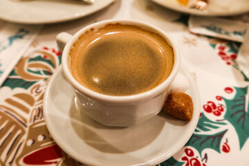 close up of hot black coffee with foam on top with a brown sugar on the side on a table cloth with Christmas red and green pattern