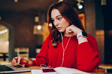 Pensive hipster girl preparing for exams searching information on netbook and listening music on cellphone for concentration,clever female student learning language via apps on smartphone and laptop