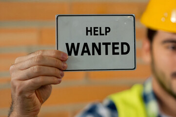 Construction worker holding a sign with the text 'help wanted'. Symbol for a job offer at a construction site.