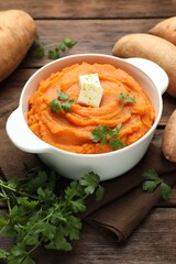 Tasty mashed sweet potato with butter in bowl, fresh vegetables and parsley on wooden table, closeup