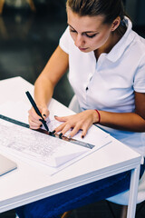 Professional female architect drafting blueprint accountings using ruler at desktop with netbook,...