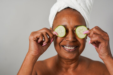 Happy senior african woman holding cucumber slices in front of her eyes during beauty treatment -...