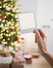 Polaroid mockup in hand over christmas interior with christmas tree, stars, gifts and decoration. Winter holiday xmas home decor with instant photo frame mock up