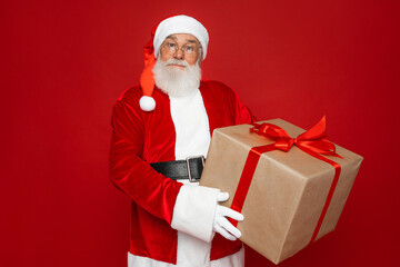 Portrait of his he nice handsome attractive cheerful overweight glad bearded Santa father holding in hands big large craft giftbox, celebrate isolated bright vivid shine vibrant red color background