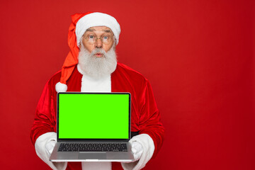 Surprised Santa Claus man in Christmas hat red coat holding using laptop pc computer showing demonstrate blank empty screen isolated on red background copy empty space