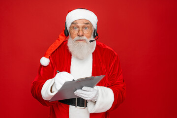 Amazed mature Santa Claus with real beard in red hat headset holding pen and clipboard, isolated on...