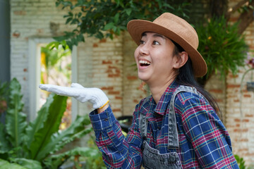 Asian woman in plaid shirt, denim overalls, and straw hat smiling with playful gesture wearing white gloves in backyard garden with vibrant plants and rustic setting.