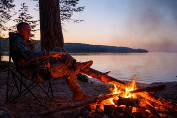 A man sits in a camping chair by a forest lake at dusk, warming his hands by a crackling campfire. He gazes into the flames, enveloped in the peaceful ambiance of nature and camping bliss.