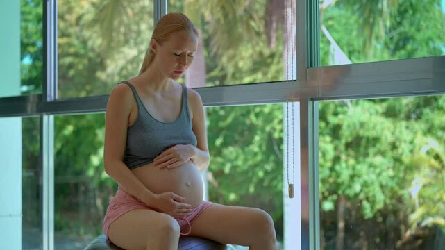 A pregnant woman sits on a fitness ball in front of a large sunlit window, gently swaying and practicing breathing techniques to release tension during contractions. The scene radiates calmness and