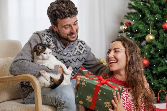 Happy couple receiving Christmas gifts in the company of their pet. Young couple give each other presents with their cat in their arms. Gifts for everyone.