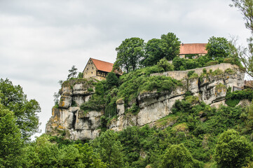 Fototapeta premium Historische Fachwerkgebäude einer Burg an steil abfallendem Felsmassiv mit Bäumen und Sträuchern vor bewölktem Himmel
