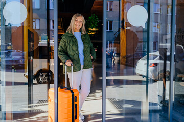 Elderly woman with blonde hair standing at hotel entrance with bright orange suitcase. Reflective...
