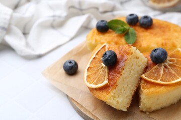 Pieces of delicious semolina cake with blueberries and orange slices on white tiled table, closeup. Space for text