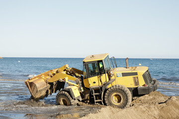 Obraz premium Yellow tractor on the beach, with sand and sea, used to smooth sand and manage erosion problems by replacing lost sand.