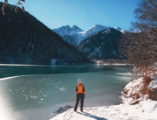 A woman tourist admires a beautiful winter lake in the mountains, surrounded by a spruce forest...
