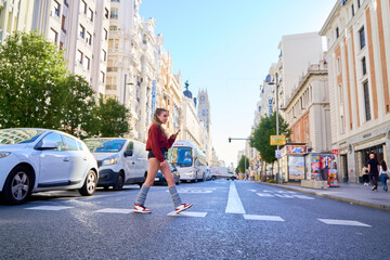 Portrait of a young Caucasian woman holding her mobile phone as she crosses a street at a pedestrian crossing.