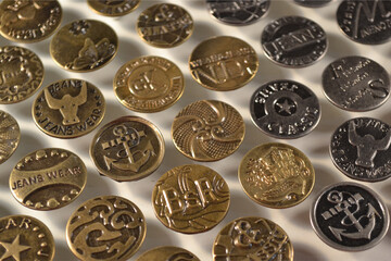 Decorative metal buttons for denim jeans, arranged neatly on white. Buttons close-up view in gold, bronze, and silver finishes, showcasing intricate engravings and stylish motifs.