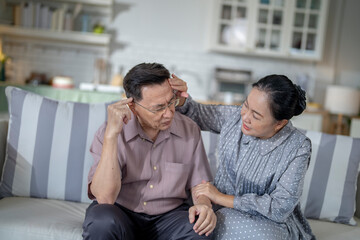Obraz premium An elderly couple sits on a couch in a cozy living room. The man holds his head in discomfort, while his partner offers support. The image highlights care, concern, and the bond between the couple.