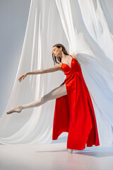 Ballerina in red long dress dancing on pointe shoes in white light studio with white cloth