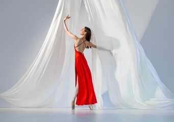 Ballerina in red long dress dancing on pointe shoes in white light studio with white cloth