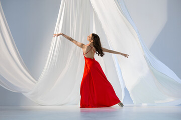 Ballerina in red long dress dancing on pointe shoes in white light studio with white cloth