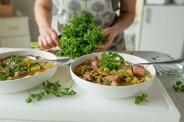 Woman with apron serving a fresh homemade cooked green bean soup with meat in the kitchen