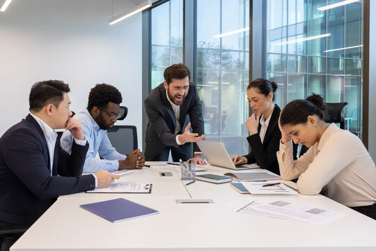 Angry boss yelling at colleagues during business meeting in modern office. Diverse team displays stress and frustration while discussing work issues around conference table.