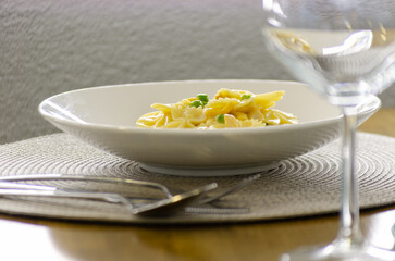 A bowl of creamy pasta with peas, served on a placemat with a wine glass in the background. The image is a close-up of the pasta, highlighting its texture and color.
