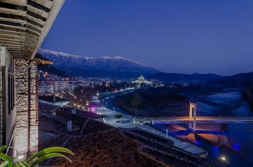 A panoramic night view of Berat in Albania with the Osumi River flowing through it. The city is illuminated with warm lights, while the snow-capped Tomori mountain in the background 