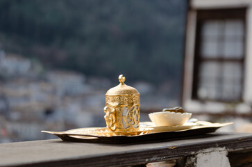 A traditional Turkish coffee set, made of gold and porcelain, rests on a wooden tray. The set is placed on a balcony overlooking the picturesque town of Berat in Albania