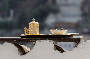 A traditional Turkish coffee set, made of gold and porcelain, rests on a wooden tray. The set is placed on a white wall with a blurred cityscape in the background.