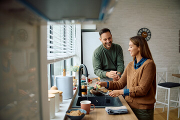 Obraz premium Happy man talking to his wife while she is washing dishes in kitchen.