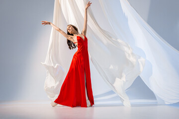 Ballerina in red long dress dancing on pointe shoes in white light studio with white cloth