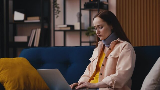 Upset woman wearing neck brace works on laptop at home, pain after accident