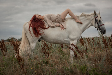 Beautiful girl model with long red hair lies on a white horse in the park at sunset