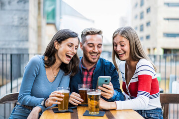 Three friends laughing together while looking at a smartphone outdoors. Young adults enjoying a fun moment with beer at a city bar