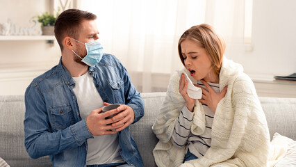 Coronavirus outbreak. Young woman coughing at home, boyfriend in protective mask pulling back from her