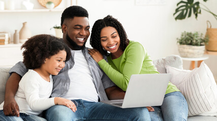 Fototapeta premium Family Weekend. Cheerful black parents and their little daughter using laptop at home together, watching movie or browsing internet, free space