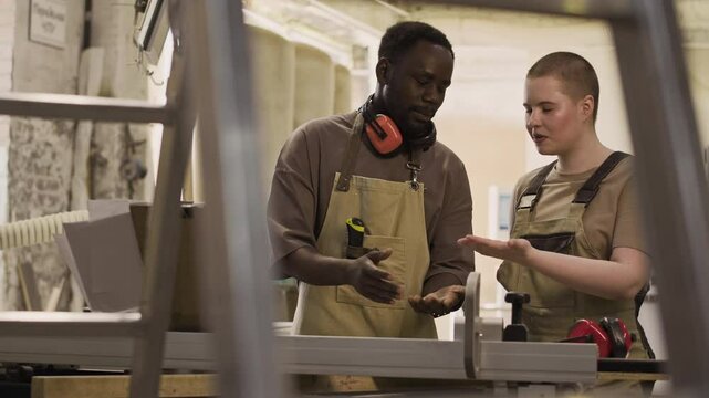 Black male woodworker, wearing apron and safety earmuffs around his neck, instructing young Caucasian female apprentice in carpentry workshop