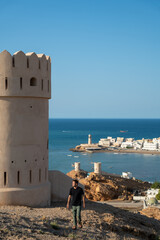 A man stands atop a historic watchtower in Sur, Oman, overlooking the surrounding coastal landscape.
