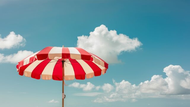 Summer Serenity: A vibrant red and white striped parasol stands open against a backdrop of a clear blue sky dotted with fluffy white clouds, evoking feelings of relaxation and leisure.