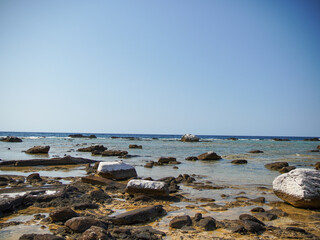 A tranquil beach scene showcases a rocky shoreline bathed in sunlight. Numerous dark rocks and several light-colored, possibly bleached, stones are scattered across the shallow water.
