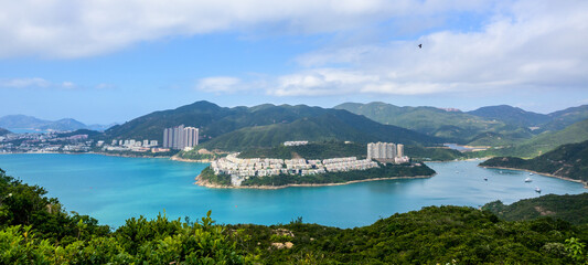 A panoramic view overlooking a bay with a peninsula featuring buildings, lush green hills, and a few boats on the water from Dragons Back Hike, Shek O Peak, Hong Kong.