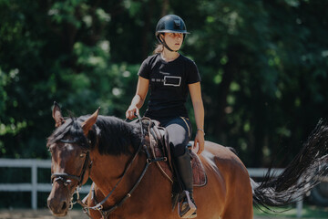 A young female equestrian rides her horse in an outdoor arena. She wears a helmet and focuses intently, surrounded by greenery, showcasing the connection between rider and horse.