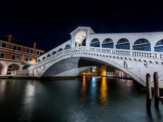 The Rialto Bridge, an iconic historical structure of Venice, is beautifully lit against the dark evening sky reflecting on the calm waters of the Grand Canal.