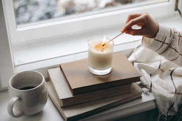 Kids hand in checkered pyjamas with lighting candle on windowsill. Autumn still life with cup of...