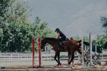 A young rider, wearing a helmet, practices horse jumping over obstacles in an outdoor arena....