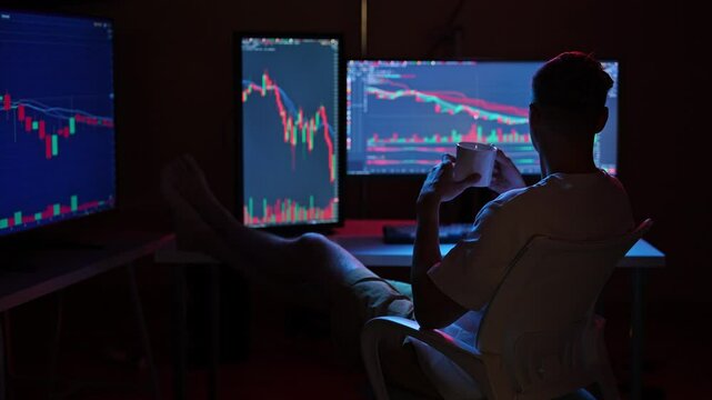 Rear view of relaxed male trader analyzing stock market charts on multiple computer screens in home office at night, sipping coffee with feet up on desk. Concept of online trading and investment.