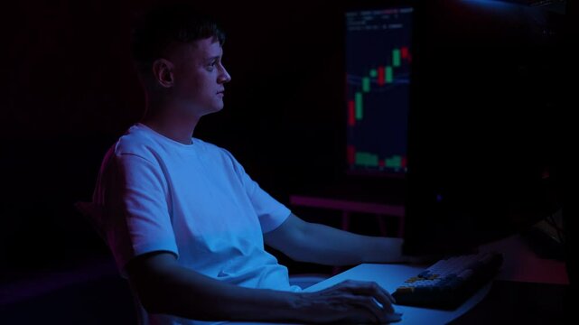 Side view of focused young male trader working late at night on multiple computer monitors, analyzing financial data and making investment decisions based on graphs and charts. Shooting in slow motion
